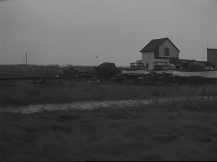 Blast Of Silence (1961) movie still: establishing — A rural boatyard on a cloudy day features several boats on cradles near a structure. Tall grass…