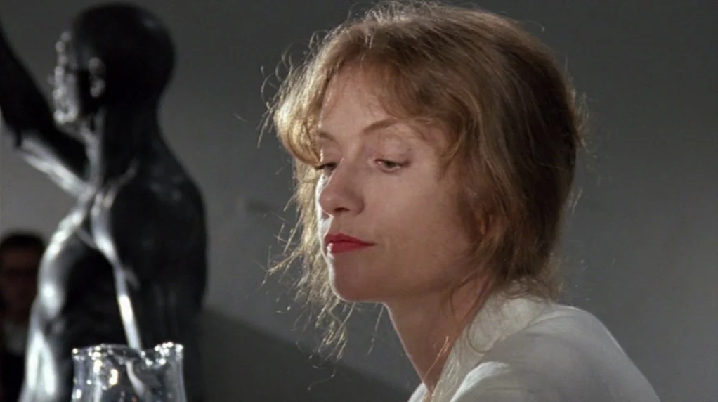 Malina (1991) movie still: closeup — An interior space displays a dark anatomical sculpture in the background. A woman in a white shirt looks…
