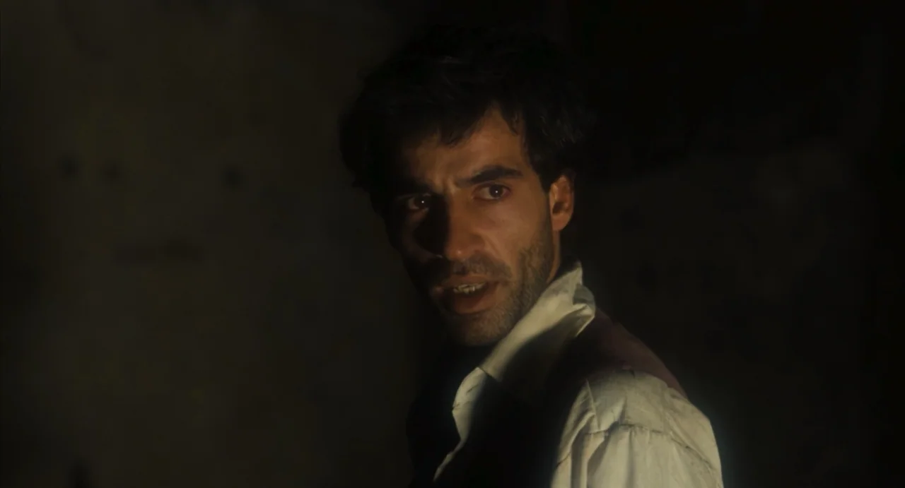 Camila (1984) movie still: closeup — A dimly lit room with heavy shadows obscuring the background. A man looks off-camera while illuminated by a…
