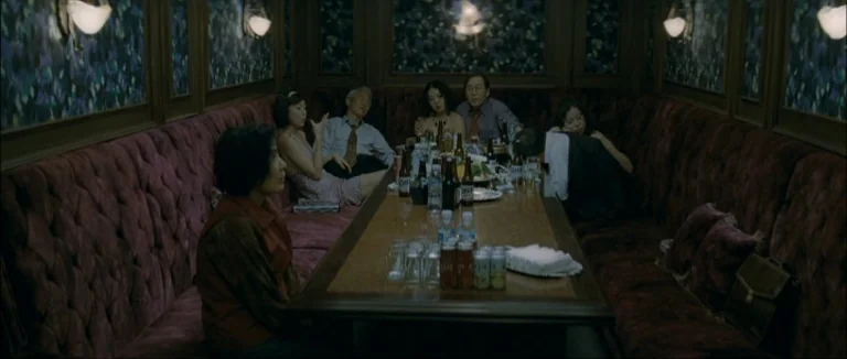 Mother (2009) movie still: wide — A dimly lit private karaoke room features plush burgundy tufted booths and patterned wallpaper. Several adults sit…