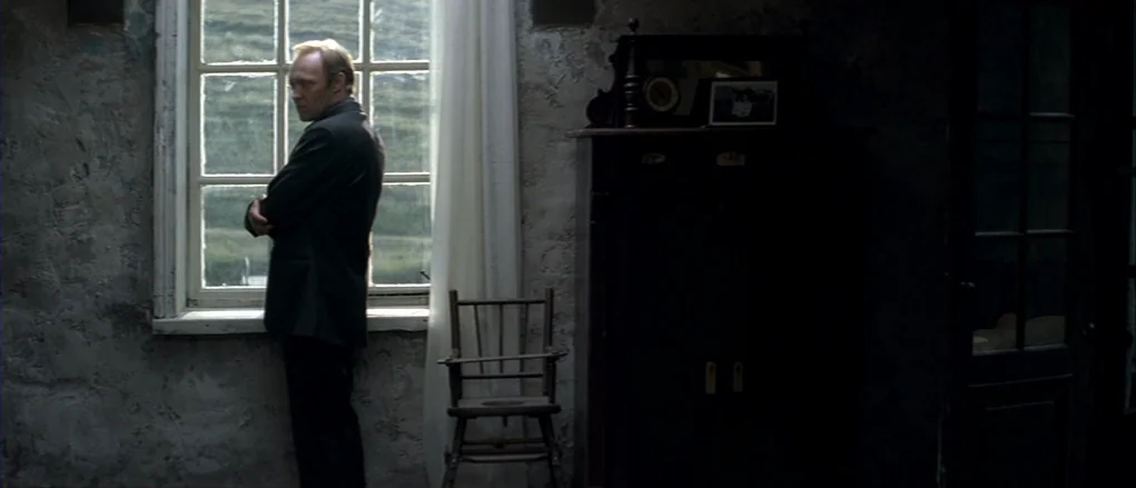The Banishment (2007) movie still: medium — A dimly lit room with distressed plaster walls and an old-fashioned window pane. A man stands beside the…