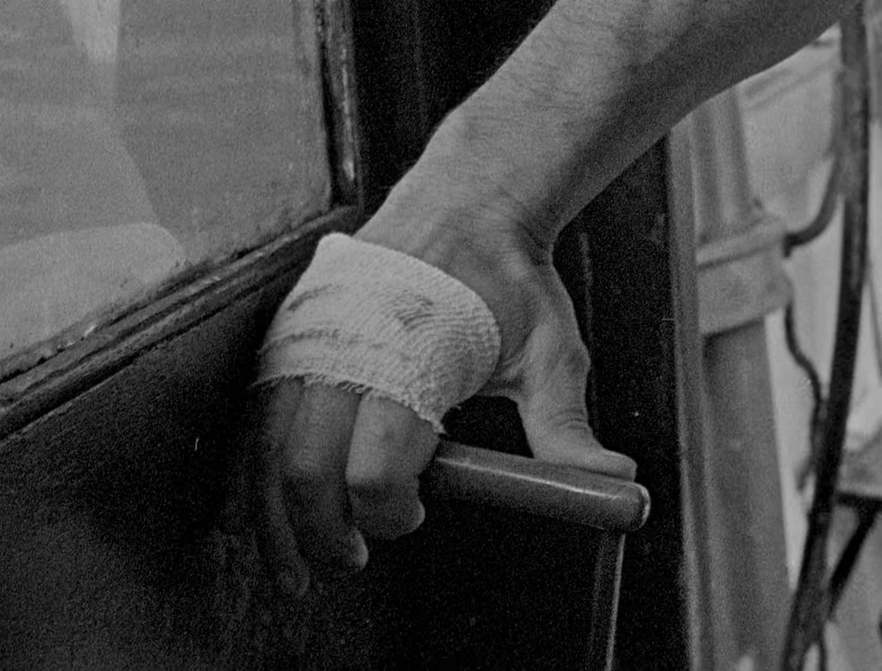 Bait (2019) movie still: closeup — A close-up view shows a hand with a white bandage on its back. The hand is gripping the handle of a dark-framed…
