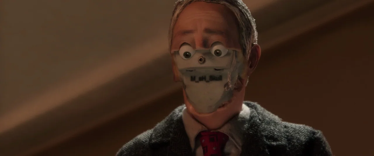 Anomalisa (2015) movie still: closeup — A stop-motion puppet with a visible mechanical lower face plate stands against a dark wall. The figure wears…