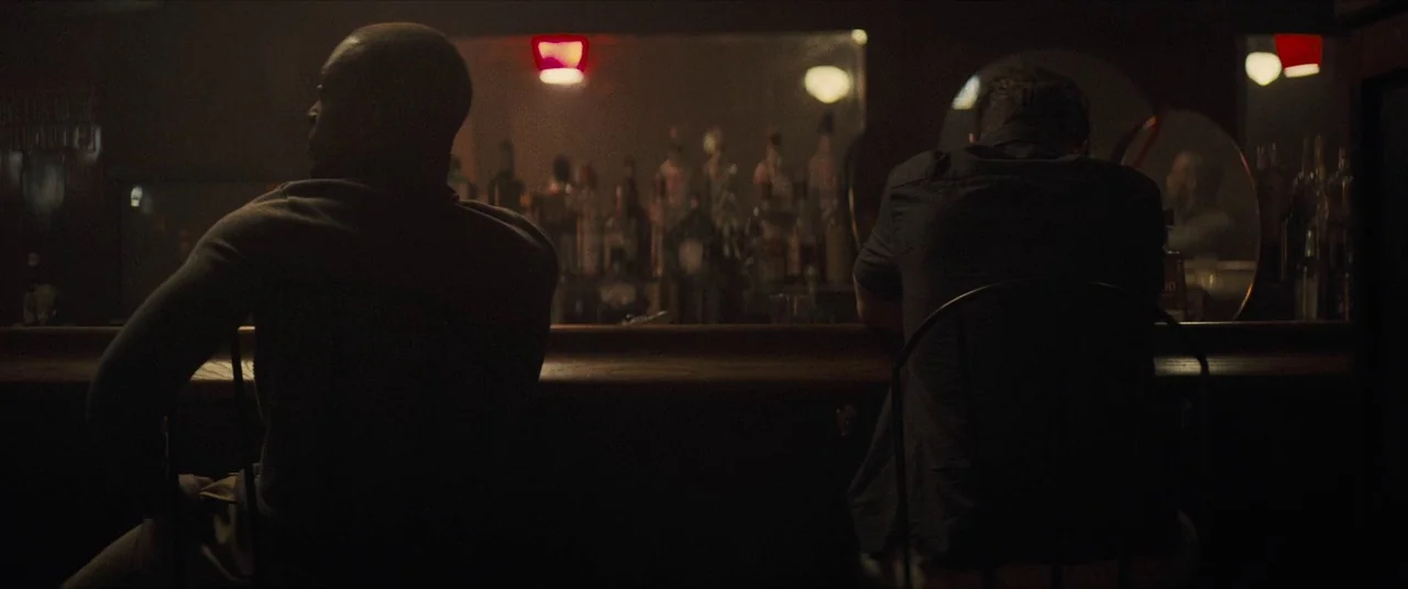 Synchronic (2019) movie still: medium — A dimly lit tavern with a wooden bar and glowing red exit signs. Two men sit at the bar counter with their…