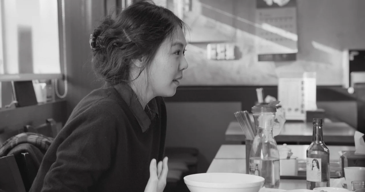 The Day After movie still: medium — A restaurant interior features tables with glassware and condiments in a simple setting. A young woman sits at a…