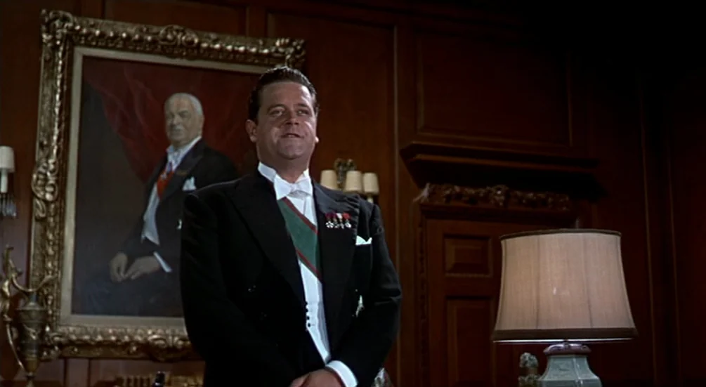 The Man Who Knew Too Much (1956) movie still: medium — A wood-paneled room features a large gold-framed oil painting on the left wall. A man in…