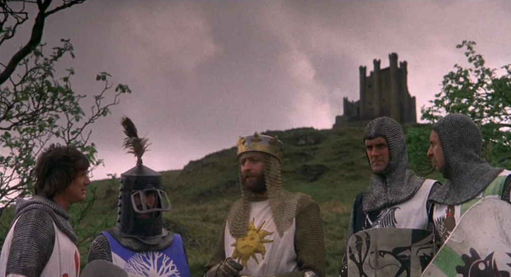 Monty Python and the Holy Grail (1975) movie still: medium — A group of five men wearing medieval knight costumes stand on a grassy hillside. An old…