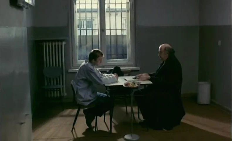 No End (1985) movie still: medium — A austere room has plain grey walls and a metal radiator against the wall. Two men sit at a small table near a…