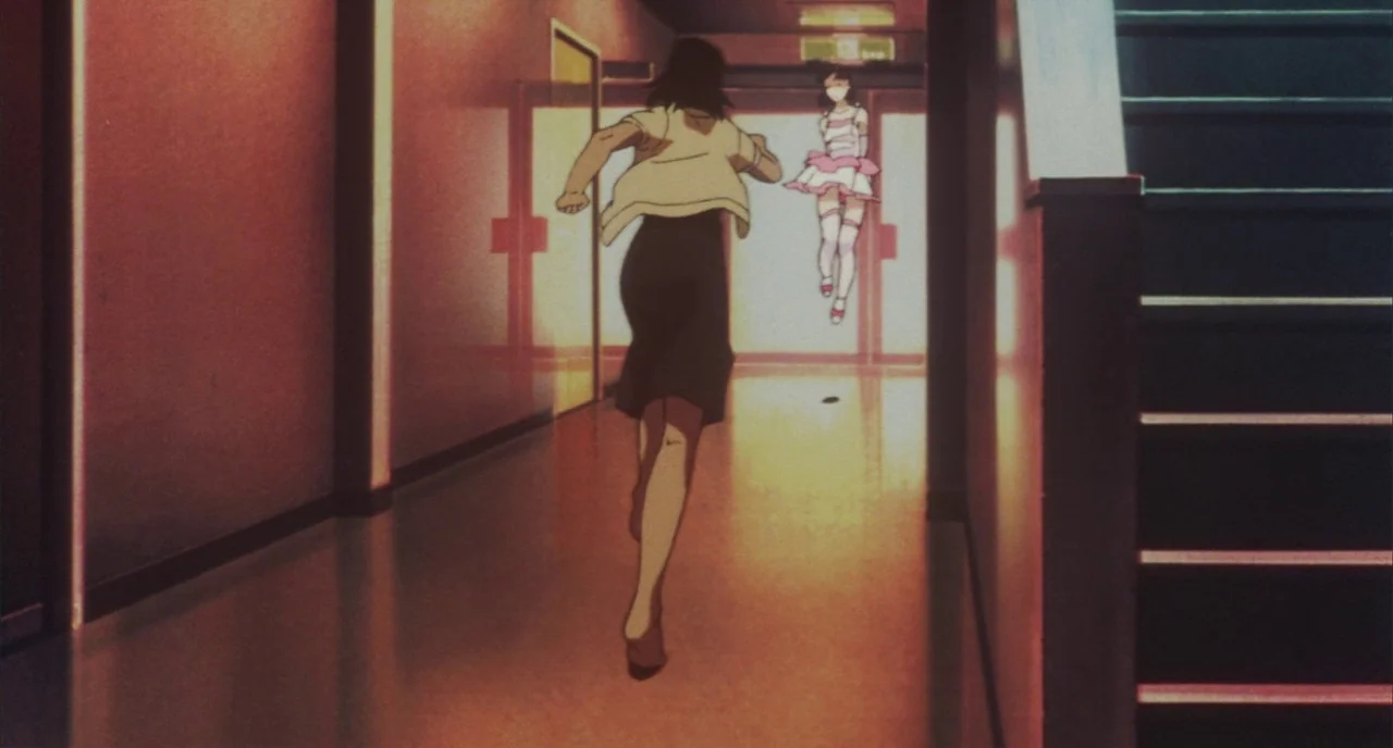 Perfect Blue (1997) movie still: fullbody — A dimly lit indoor corridor has brown walls and a staircase on the right side. A woman runs toward a…