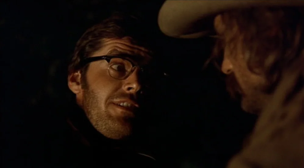 Easy Rider (1969) movie still: closeup — A dark outdoor space at night with minimal illumination on the two figures. A man with glasses looks toward…