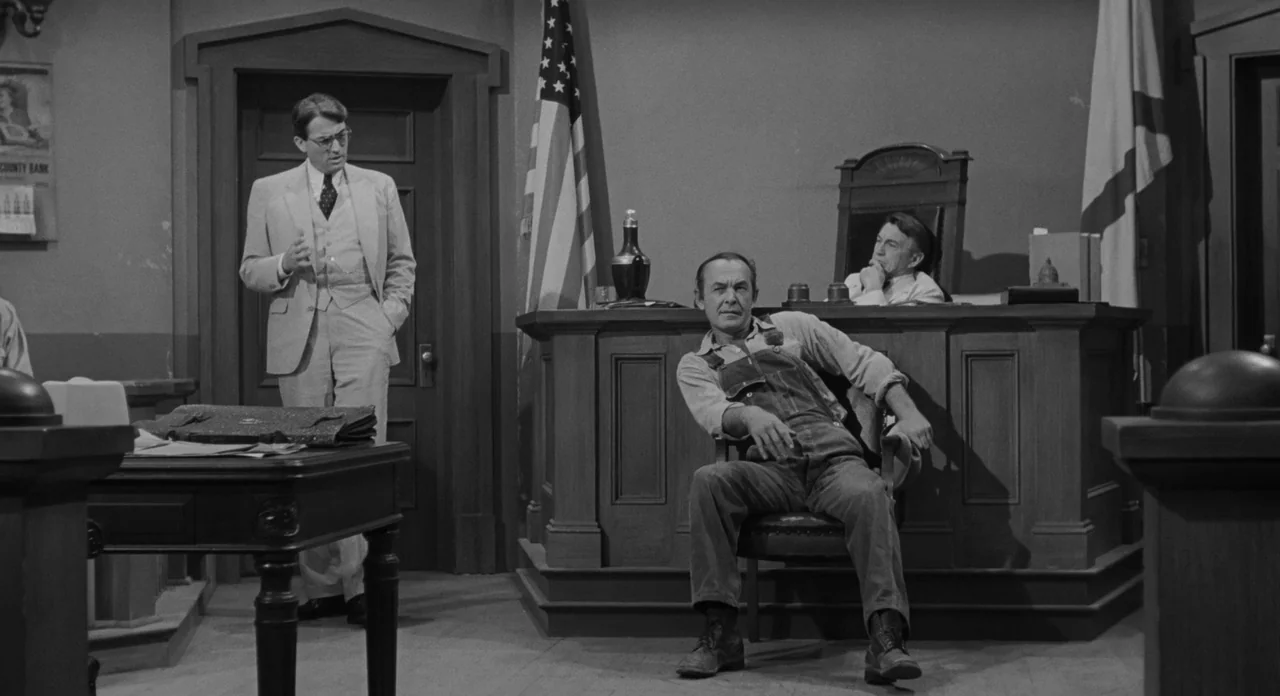 To Kill A Mockingbird (1962) movie still: medium — A vintage 1930s style wooden courtroom with a raised judge bench and large flags. A lawyer stands…