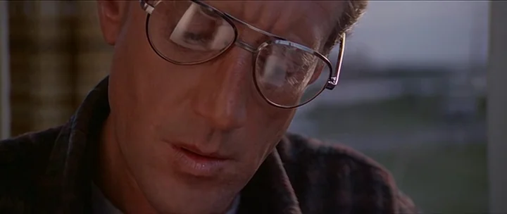 Jaws movie still: closeup — A man looks down while wearing metal-framed glasses in a brightly lit interior space. He wears a plaid shirt and appears…