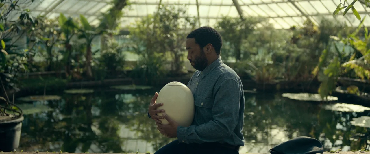 The Pod Generation (2023) movie still: medium — A glass-roofed greenhouse filled with lush tropical plants surrounds a small dark pond. A man sits…