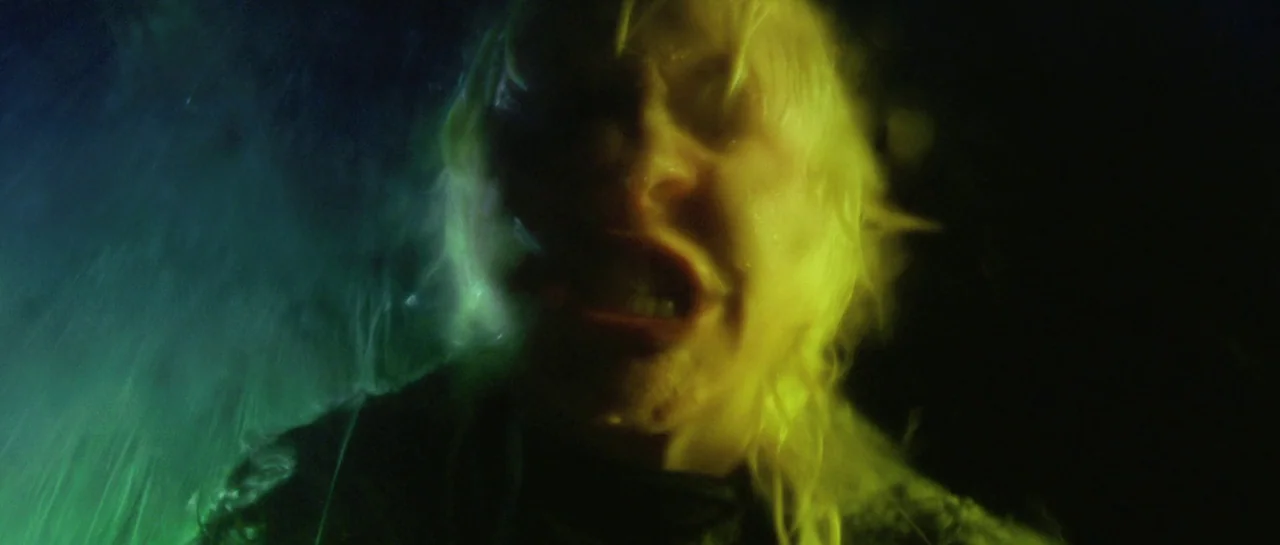 After Blue (2021) movie still: closeup — A dark, wet outdoor environment at night is illuminated by a sharp green light. A person with wet blonde…