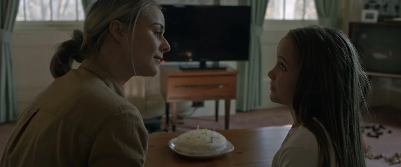 Motherly (2022) movie still: medium — A domestic interior with a birthday cake placed on a wooden surface between two people. A woman with blonde…