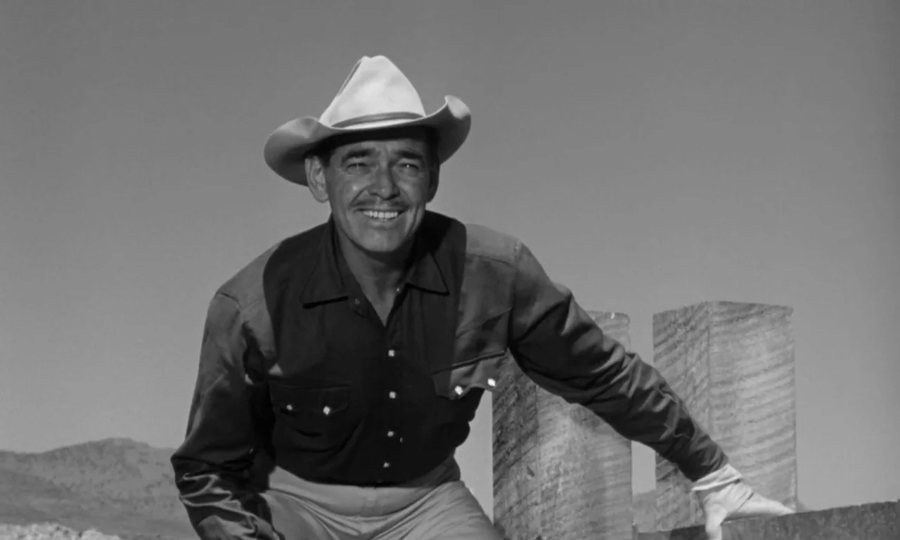The Misfits (1961) movie still: medium — An arid outdoor landscape features a man positioned near a rough-hewn wooden post. The subject smiles…