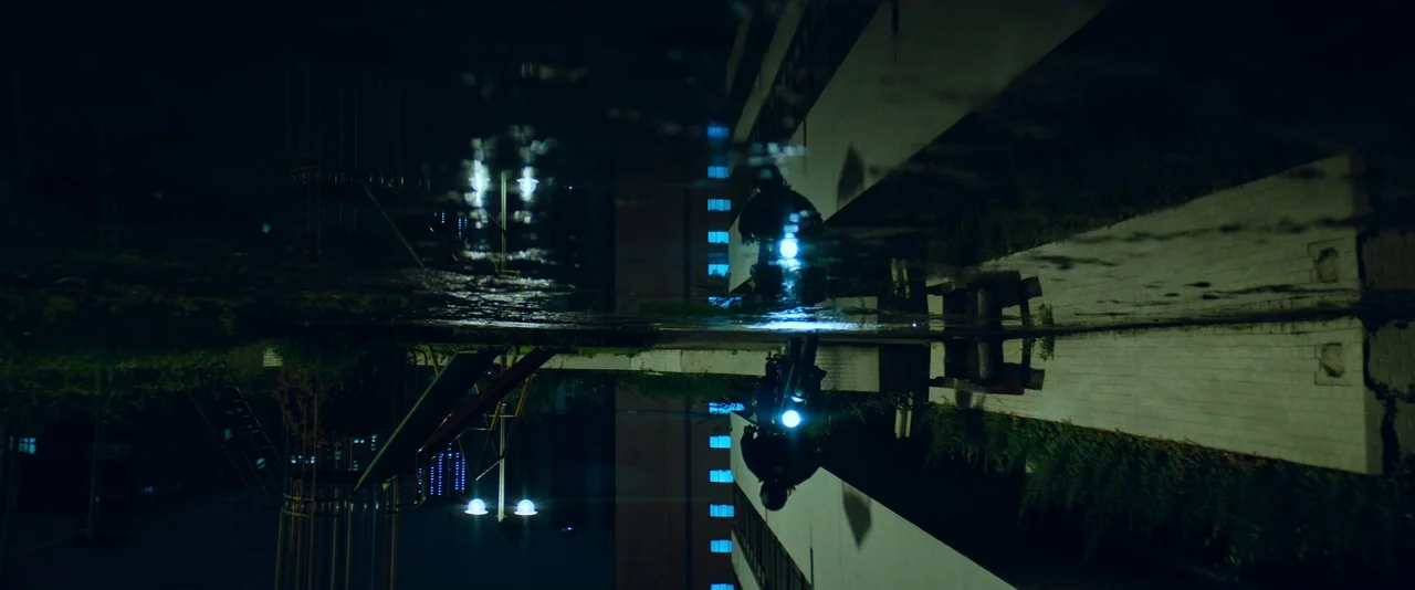 Ballerina (2023) movie still: wide — A nighttime urban scene shows a reflection in a large, dark pool of water. The inverted image reveals…
