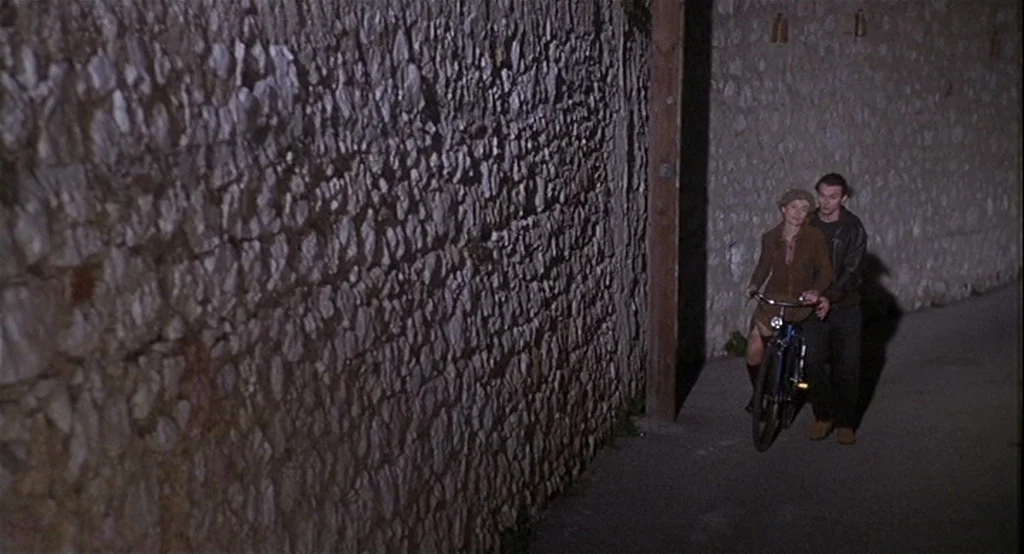 Deux (2002) movie still: wide — A narrow pathway lined with a high, rough-hewn stone wall at night. A young woman rides a bicycle as a man walks…