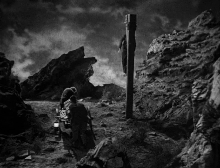 Frankenstein movie still: wide — A desolate, rocky desert terrain under a dark, cloud-filled sky. Two men stand beside a wooden gallows while a…