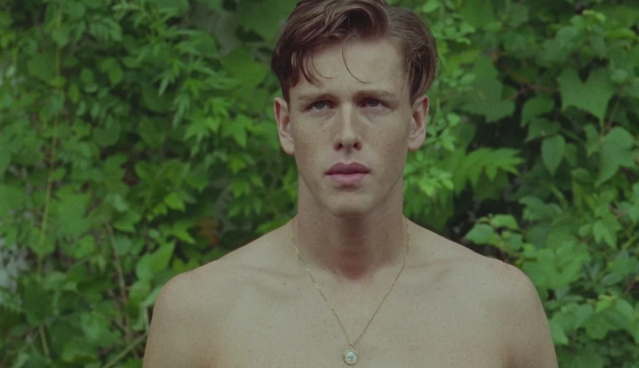 Beach Rats (2017) movie still: medium — A bare-chested young man stands outdoors against a wall of thick green leaves. He faces the camera directly…