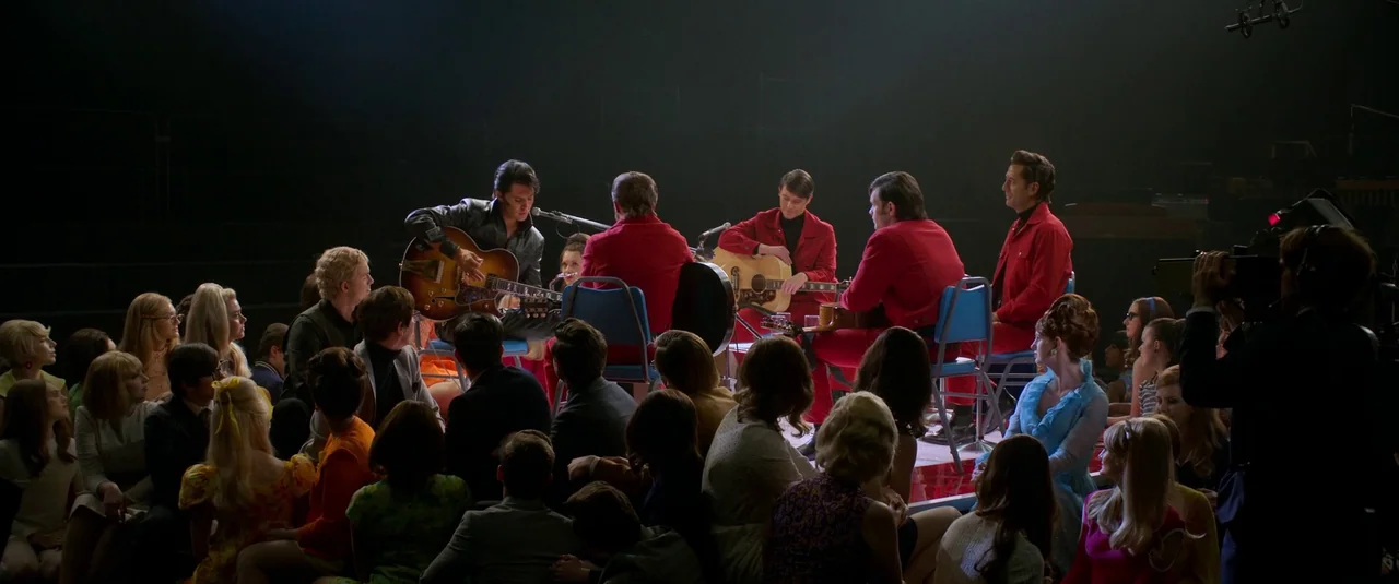 Elvis (2022) movie still: wide — A group of musicians performs in a circle on a dark, sparsely lit stage. A seated audience watches them from the…