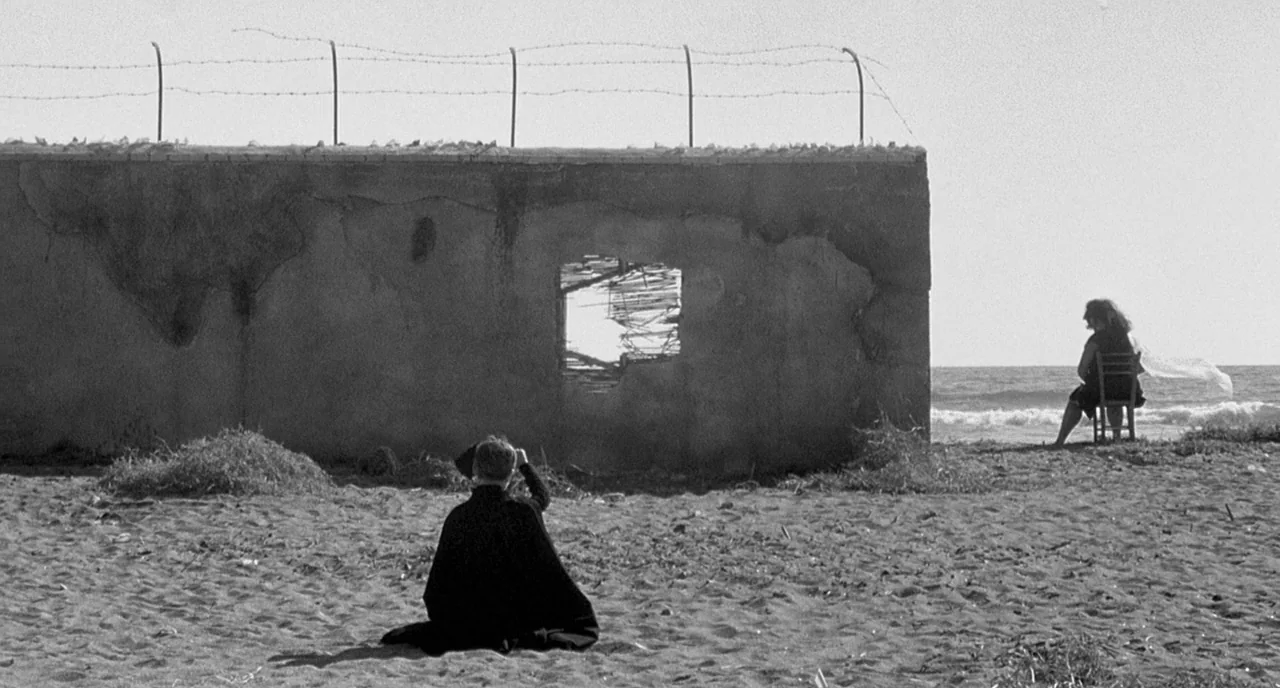 8 1/2 (1963) movie still: wide — A sandy beach features a weathered concrete wall topped with barbed wire fencing. A person sits on the sand facing…