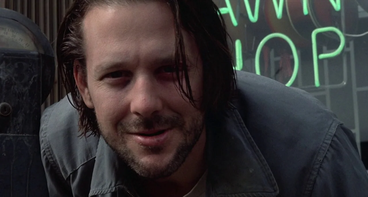 Barfly (1987) movie still: closeup — A city street at night featuring a blurred shop window with green neon light. A man leans forward to look into…