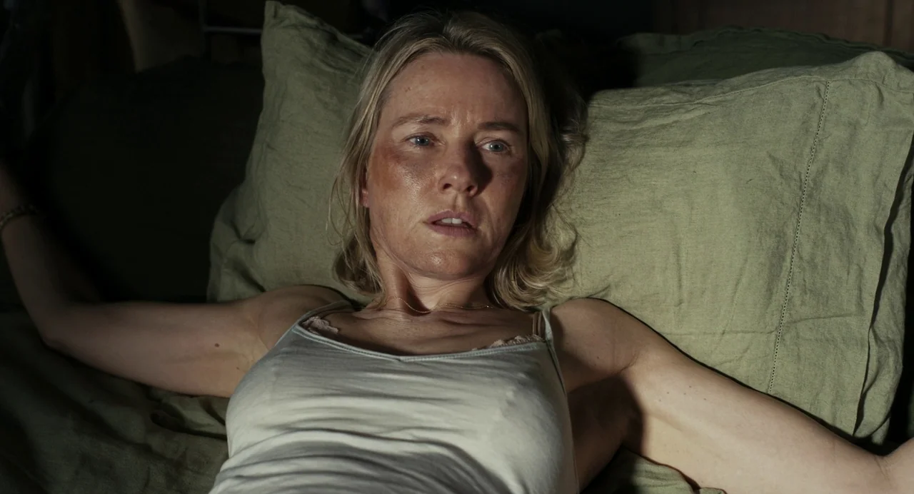 Goodnight Mommy (2022) movie still: closeup — A dark bedroom with olive green linens on the mattress and pillows. A blonde woman with visible facial…