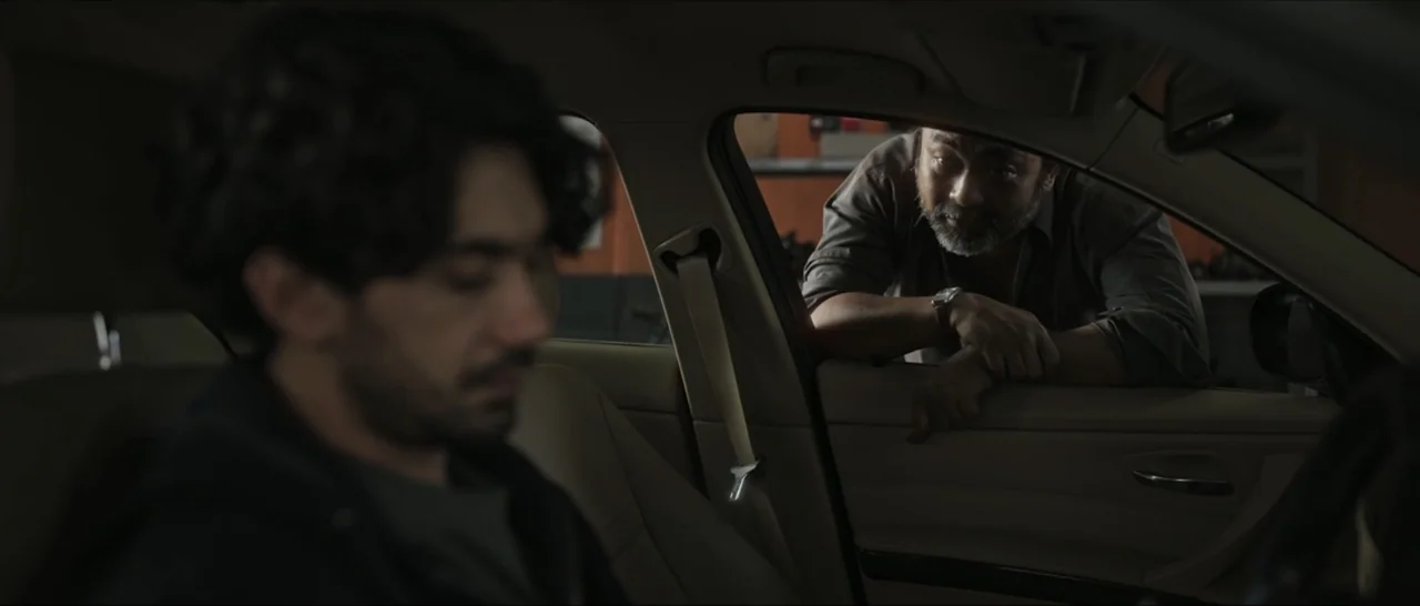 Innocent Vengeance (2023) movie still: medium — A man leans at the open driver's side window of a beige sedan. The driver sits inside listening…