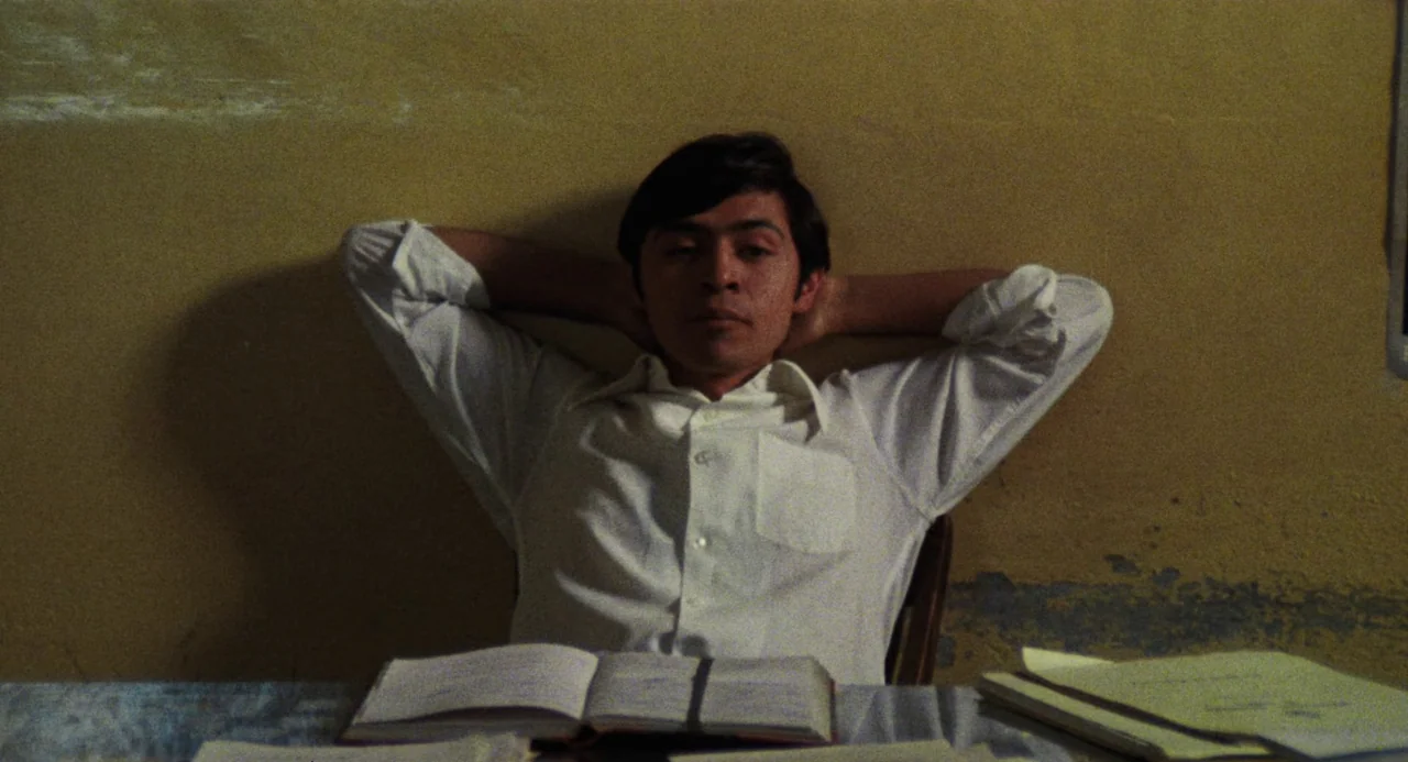 Canoa: A Shameful Memory (1976) movie still: medium — A man sits at a table against a plain, faded yellow wall. He leans back in a wooden chair with…