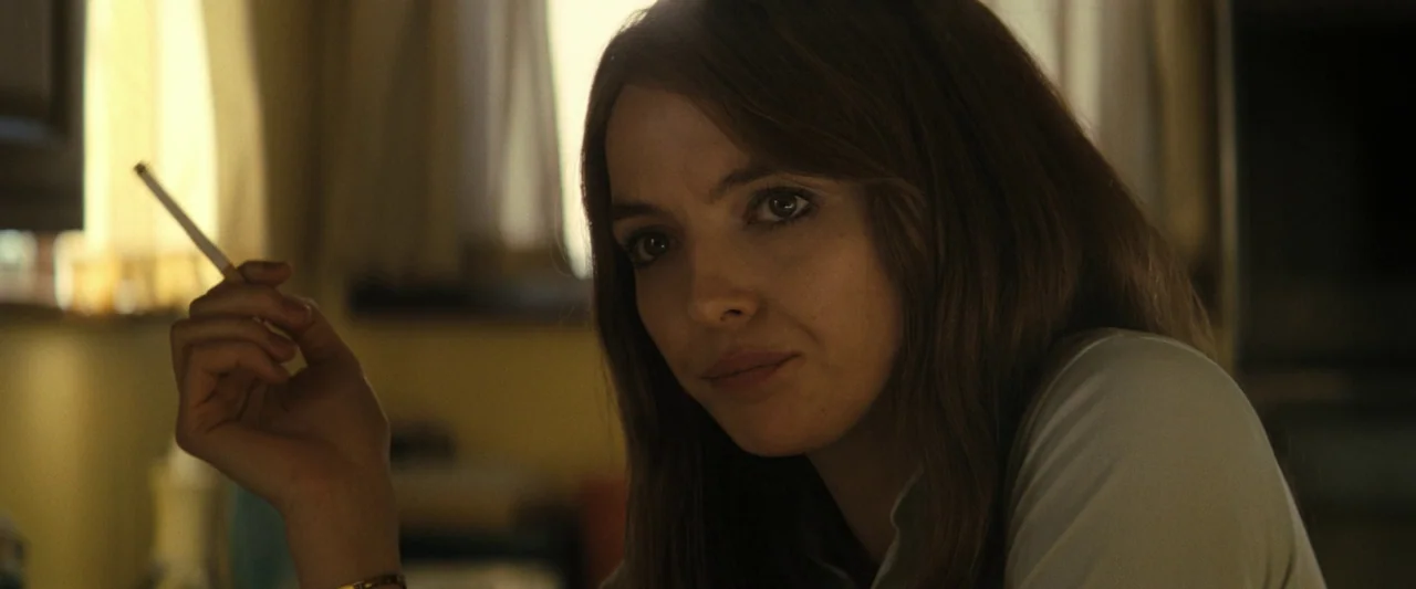 The Bikeriders (2023) movie still: closeup — An interior space with beige walls and translucent curtains visible in the background. A woman holds a…