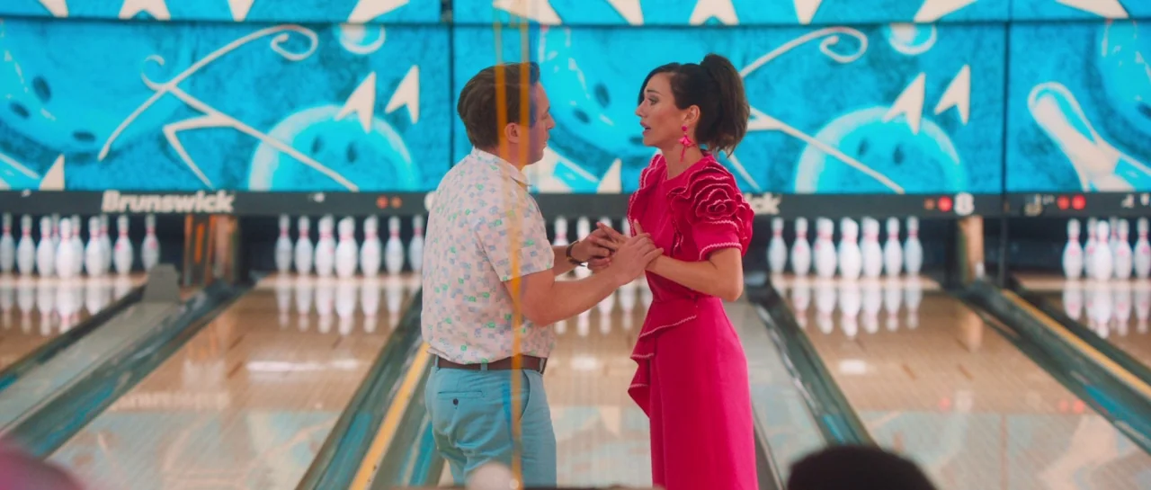 Greener Grass (2019) movie still: medium — A brightly lit indoor bowling alley with vivid blue backdrops behind the wooden lanes. A man and a woman…
