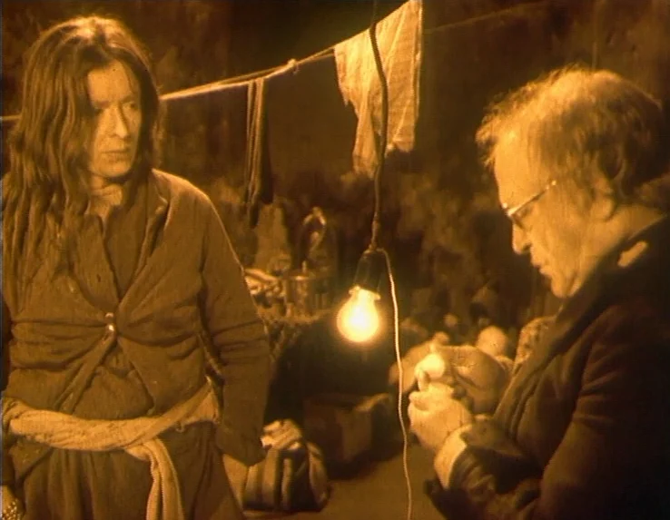 Dead Man’s Letters (1986) movie still: medium — A dark underground stone room features an exposed light bulb suspended by a cord. A woman with long…