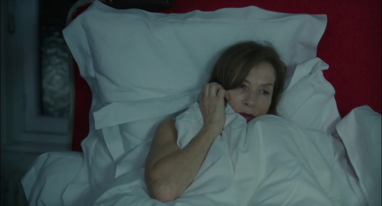 Abuse of Weakness (2013) movie still: closeup — A woman lies under a white duvet against a deep red headboard. She holds a black smartphone to her…