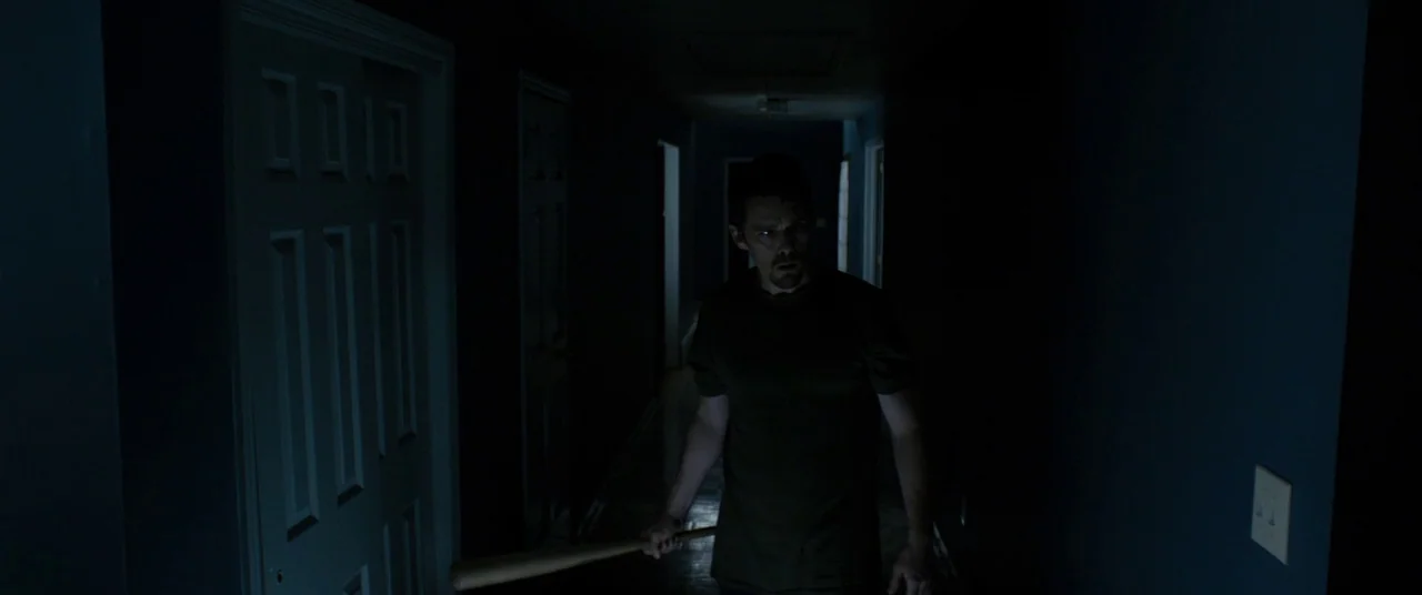Sinister (2012) movie still: medium — A dimly lit residential hallway features dark blue walls and closed white doors. A man stands in the…