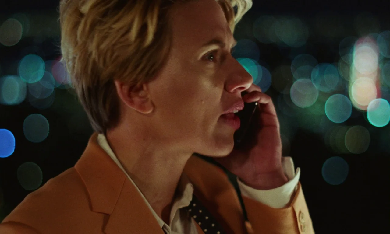 Marriage Story movie still: closeup — A woman stands on a dark rooftop at night with city lights blurred in background. She wears an orange blazer…