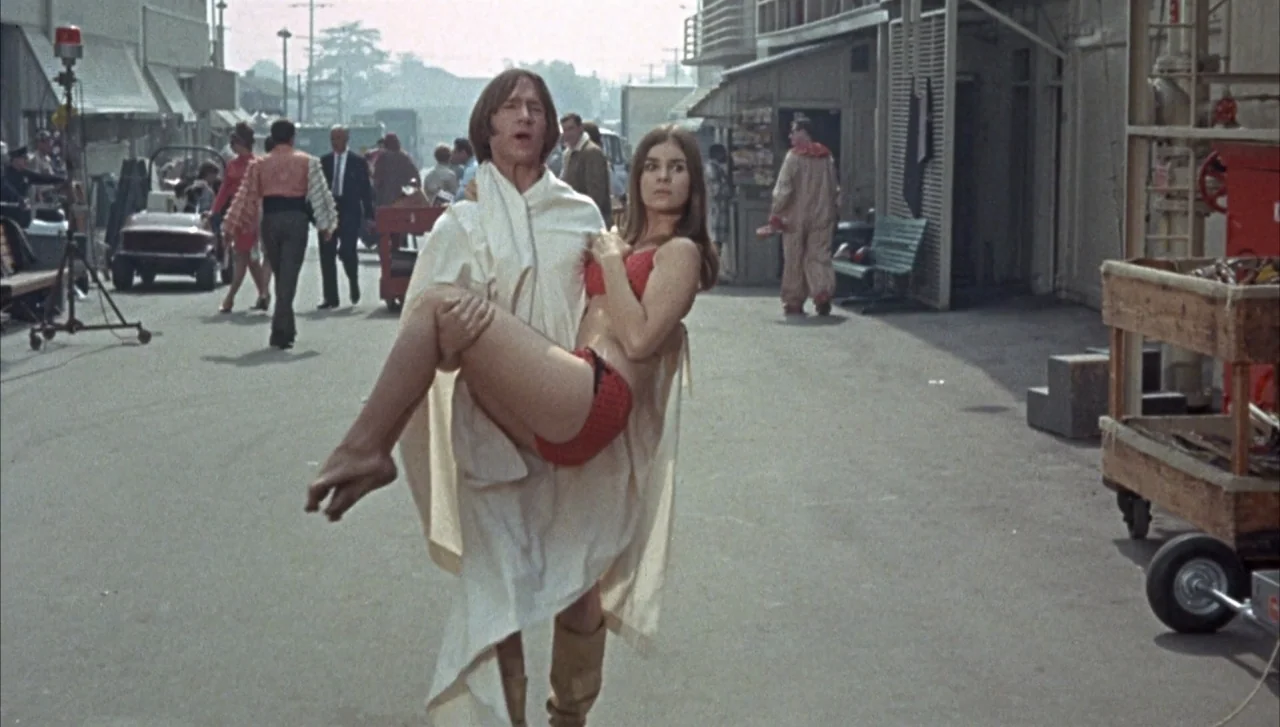 Head (1968) movie still: fullbody — A film studio backlot with various production equipment and a clown walking in background. A man carrying a…