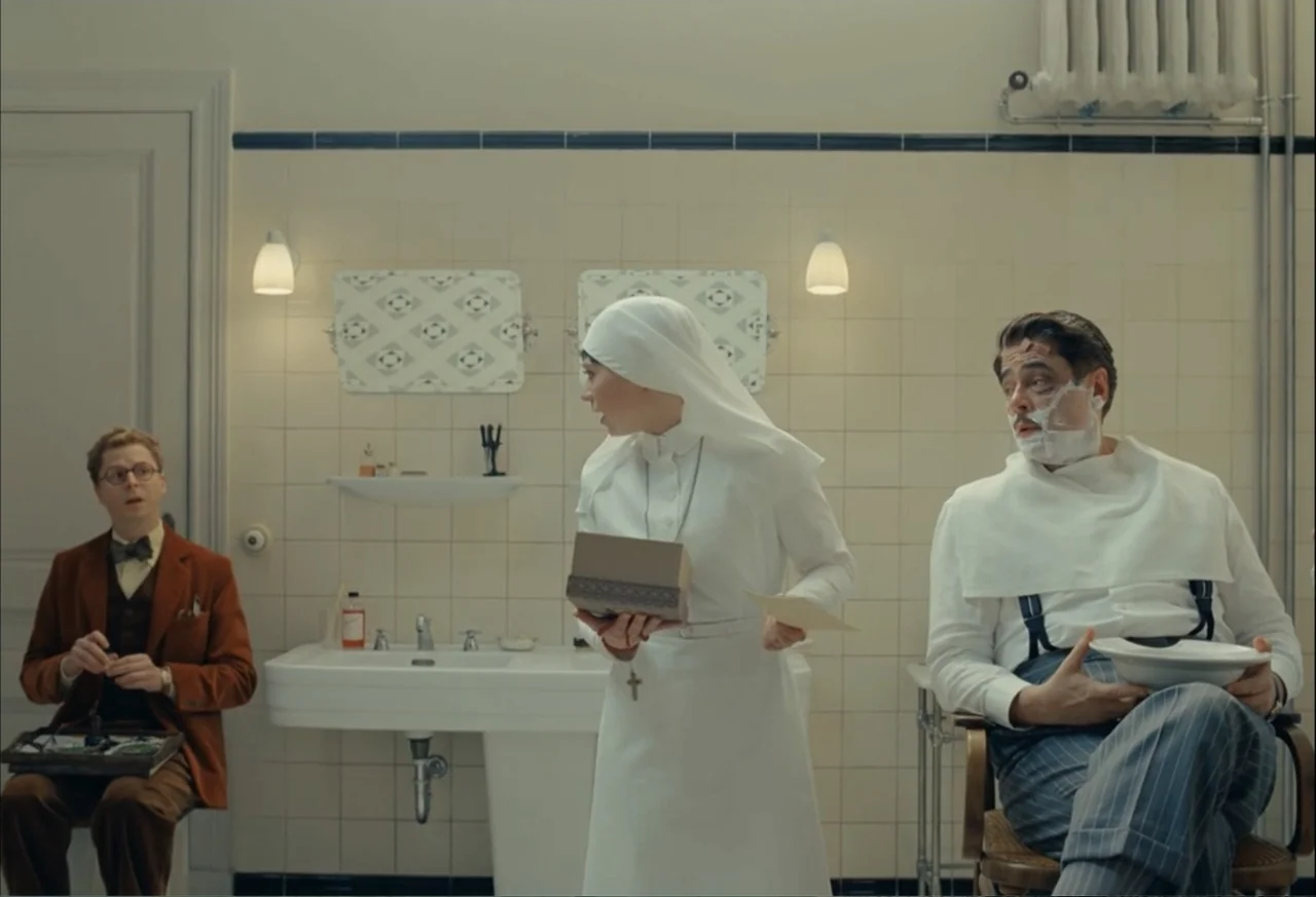 The Phoenician Scheme (2025) movie still: medium — A vintage tiled washroom features wall-mounted porcelain sinks and cream-colored wall tiles with…