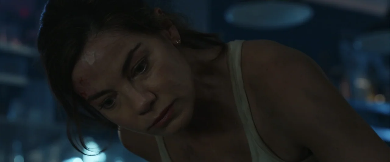 Black Site (2022) movie still: closeup — An interior dimly lit industrial space with indistinct dark surroundings. A woman with a forehead injury…