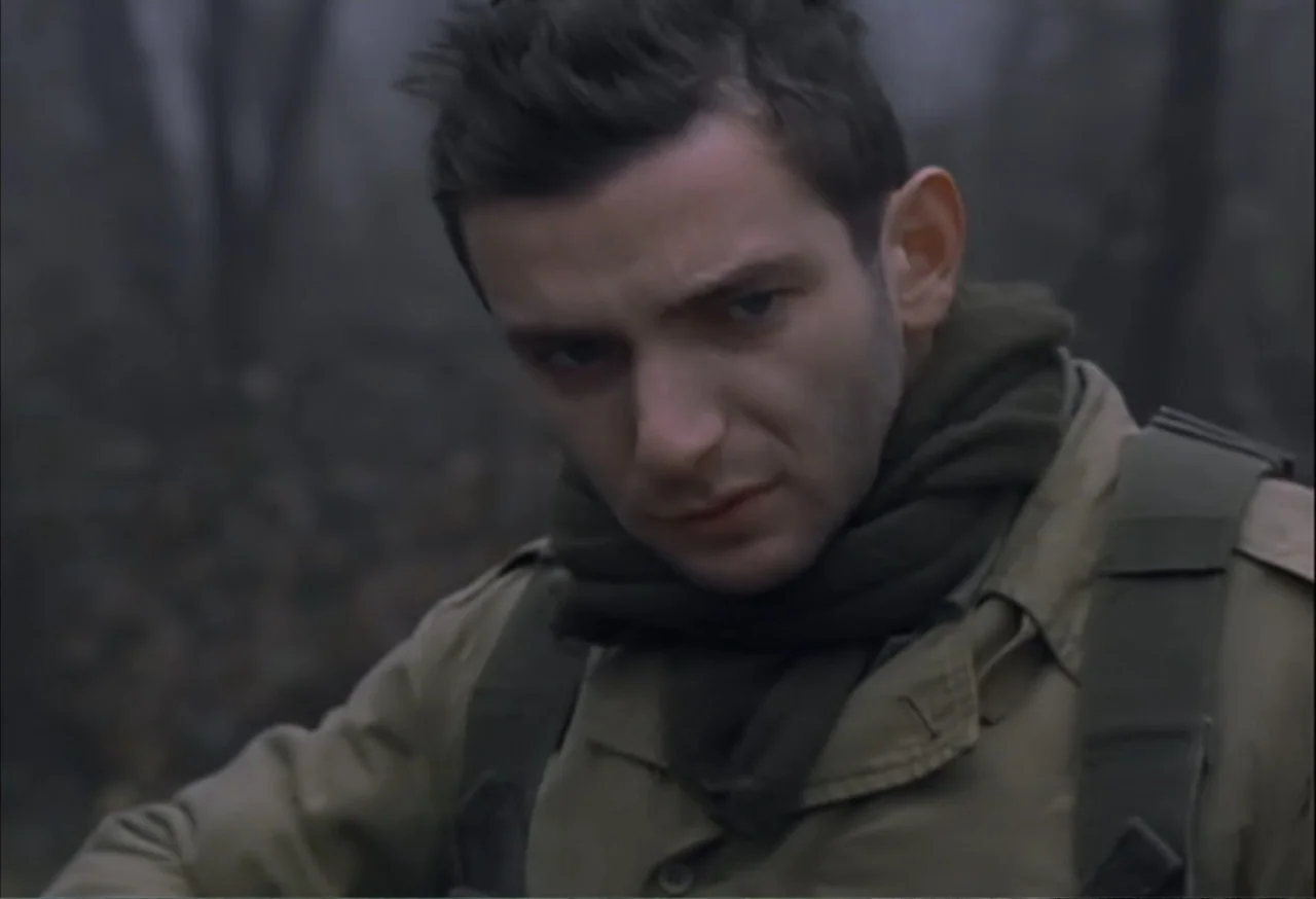 When Trumpets Fade (1998) movie still: medium — A dense, overcast woodland setting with out-of-focus trees in the background. A young man wearing…