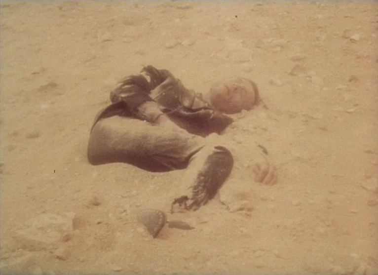 A Visitor to a Museum (1989) movie still: medium — An expanse of arid desert sand stretches across the frame. A man lies partially buried in the…