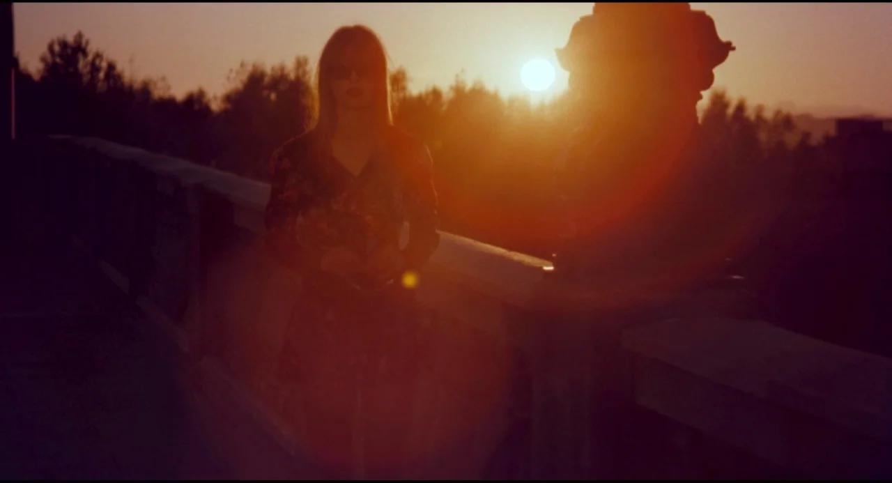 Olivia (1983) movie still: medium — A woman stands on a concrete bridge during a vibrant orange golden hour sunset. She faces the camera directly in…