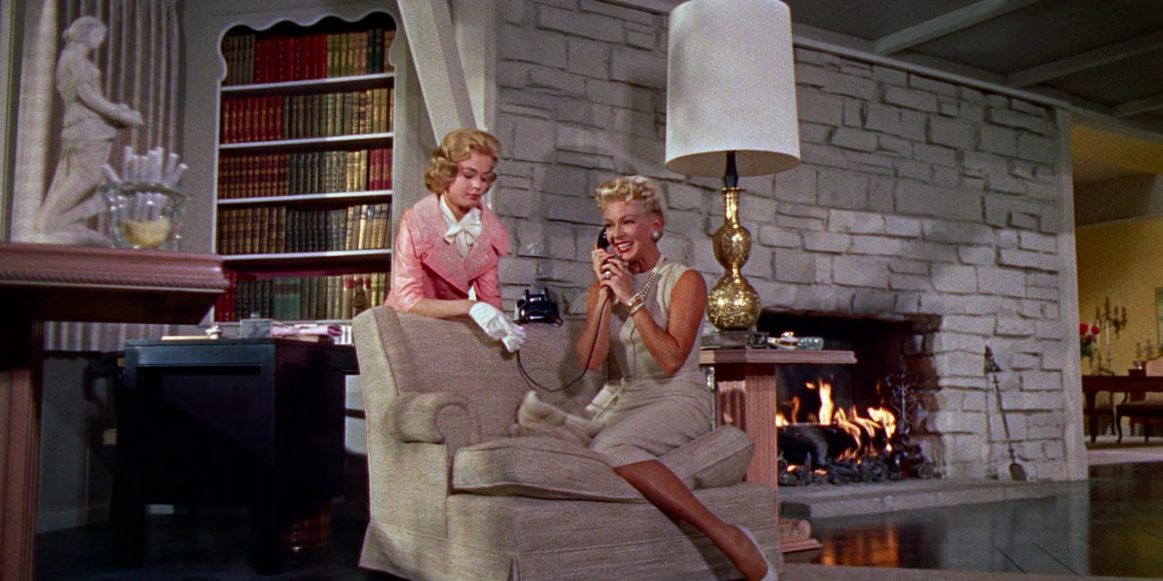 Imitation of Life (1959) movie still: medium — A 1950s living room features a stone fireplace with a fire burning inside. One woman sits in an…