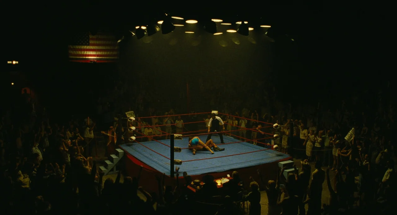 The Iron Claw (2023) movie still: wide — A dimly lit indoor arena features a blue wrestling mat surrounded by red ropes. Two wrestlers grapple in…