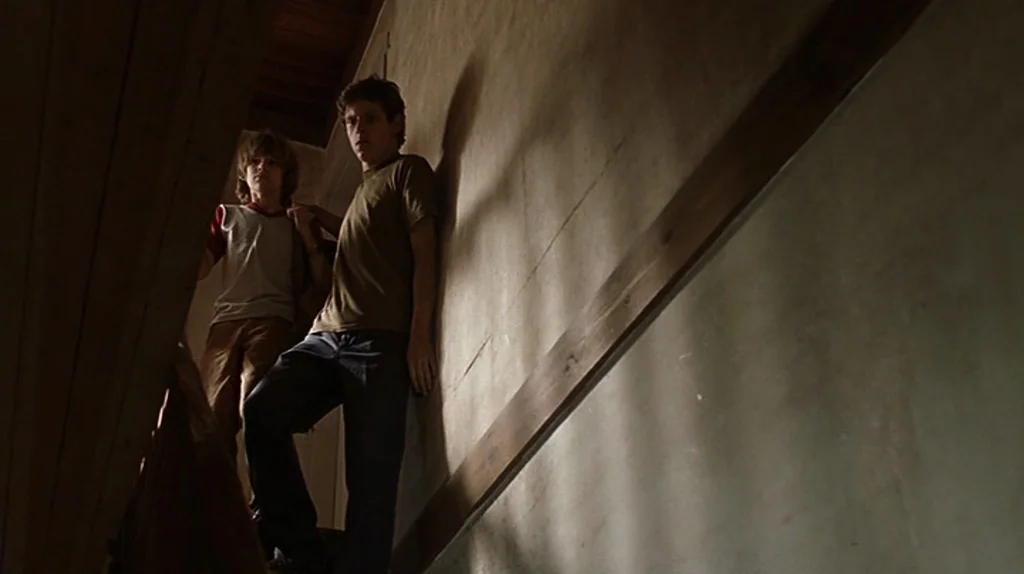 Undertow (2004) movie still: medium — A narrow residential staircase with plain plaster walls and wooden trim. Two boys stand together in the…