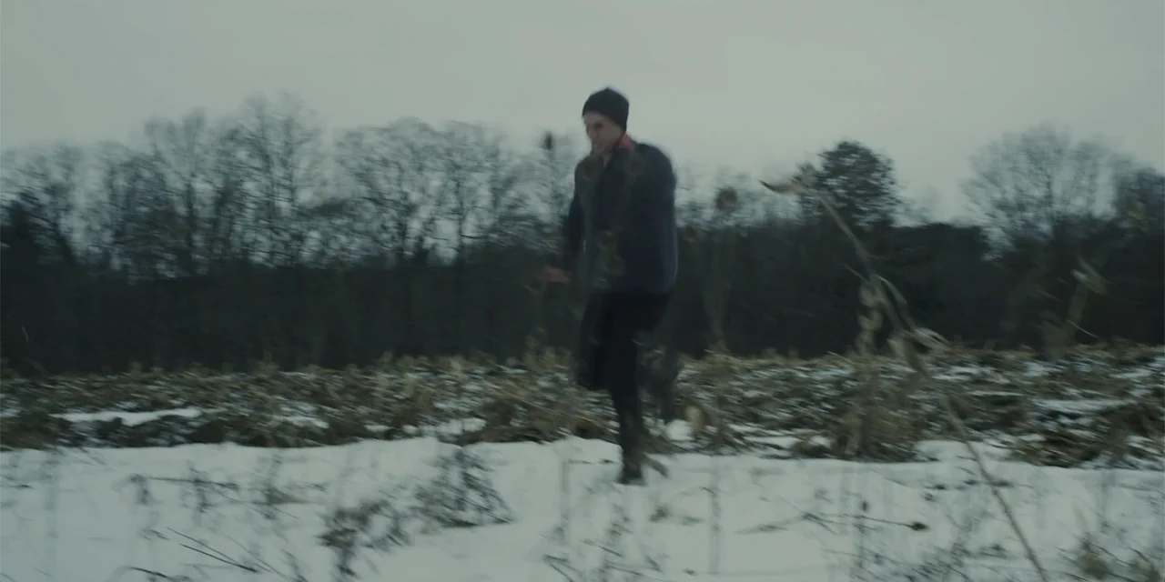 Lobos (2019) movie still: fullbody — A desolate snow-covered agricultural field lies beneath a flat grey overcast sky. A person wearing dark…