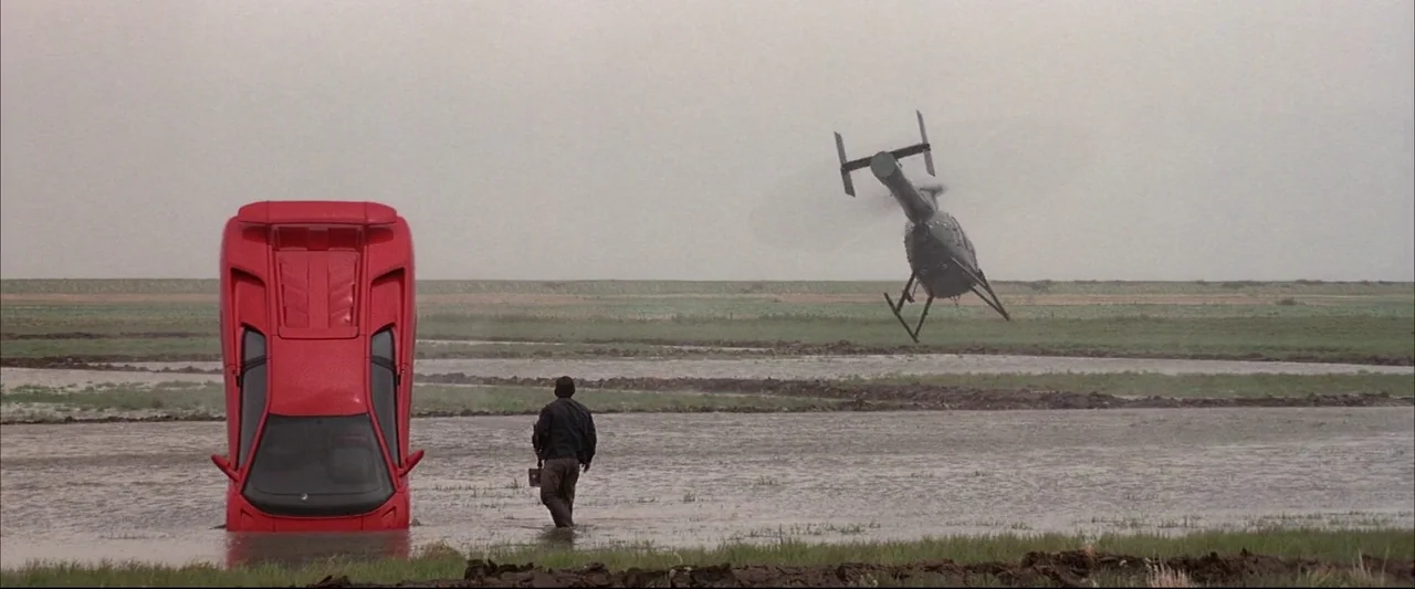 Die Another Day (2002) movie still: wide — An open flooded field under a flat grey sky contains a partially submerged vehicle. A man walks through…