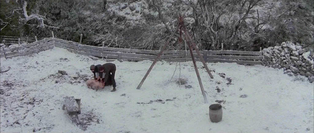 Jude (1996) movie still: wide — A snow-covered farmyard enclosed by a wooden fence and stone walls. Two figures kneel on the ground beside a pig in…