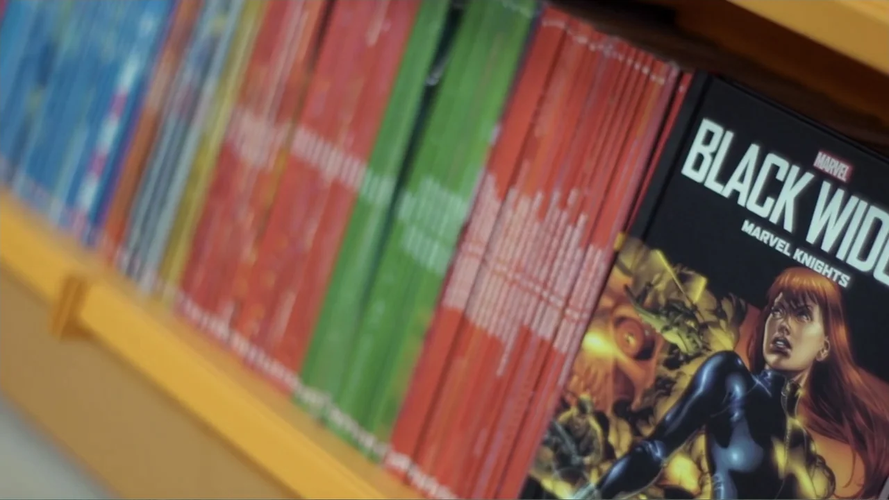 Reign of the Superwomen (2021) movie still: closeup — A close-up view of multiple graphic novels lined up on a yellow wooden shelf. The focus rests…