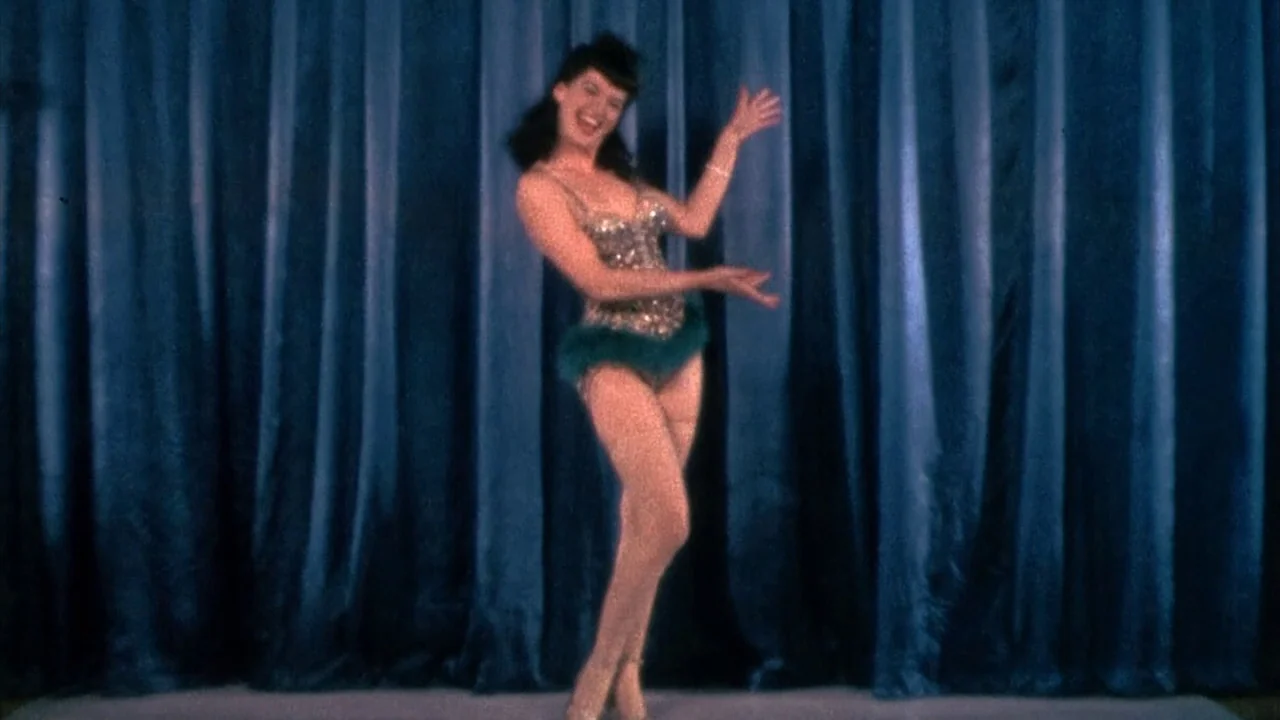 The Notorious Bettie Page (2005) movie still: fullbody — A woman stands centered in front of heavy dark blue stage curtains. The performer gestures…